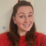 A smiling person wearing a red sweater and red hair bow, long brown hair and a small hoop earring, facing the camera; standing indoors against a light-colored door.