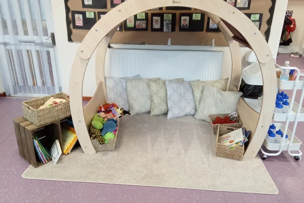 Wooden semicircular arch frames a cushioned nook holding four pillows and wicker baskets of toys and books, on a beige rug before a radiator and bulletin board in a classroom.