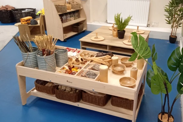 Low wooden sensory table displays organized natural play materials—bamboo sticks, wooden bowls, wicker baskets and tin containers—on a blue classroom floor with low shelves and potted plants nearby.