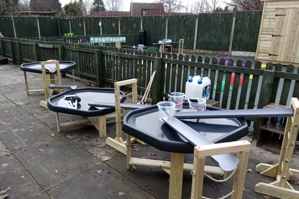 Three hexagonal plastic water play tables sit on wooden stands, each angled with channels and scoops; two clear measuring jugs and large water containers nearby in an outdoor fenced playground.