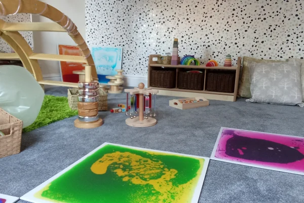 Colorful sensory mats rest on a grey carpet while wooden toys and a climbing arch are arranged nearby in a cozy children's playroom with a speckled wall and cushions.