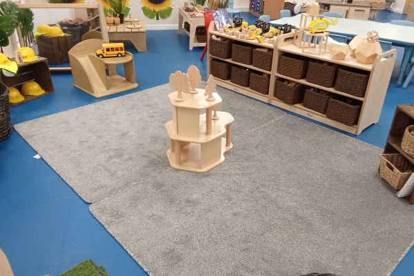 A small three-tier wooden structure with tree cutouts sits centered on two gray rugs; surrounding are wooden shelves with wicker baskets, toy vehicles, yellow safety hats, and blue flooring.
