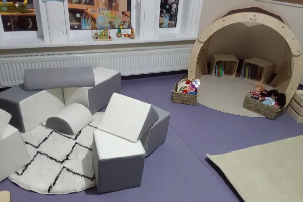 Foam modular seats arranged into a semicircular play area on purple flooring; a wooden dome reading nook holds baskets of dolls and books near a radiator and windows.