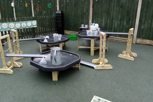 Shallow plastic basins on wooden stands, linked by angled plastic channels and holding pitchers and funnels for pouring water, arranged on a rubber playground near a green fence labeled 5 7 8 9 10.