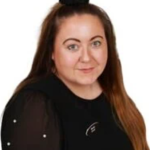 Woman faces the camera with a slight smile, long brown hair pulled into a topknot, wearing a black blouse with sheer sleeves and pearl-like studs against a plain white background.