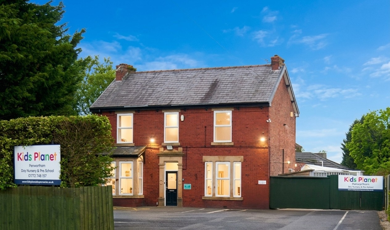 Two-story red-brick building (nursery) glowing with lit windows, standing behind a fenced forecourt and parking area, flanked by trees under a clear evening sky.